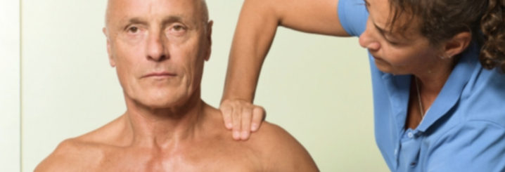 Therapeutin zeigt Grifftechnik am Schultergelenk an einem Patienten, der in die Kamera schaut. - TOP-Kurs